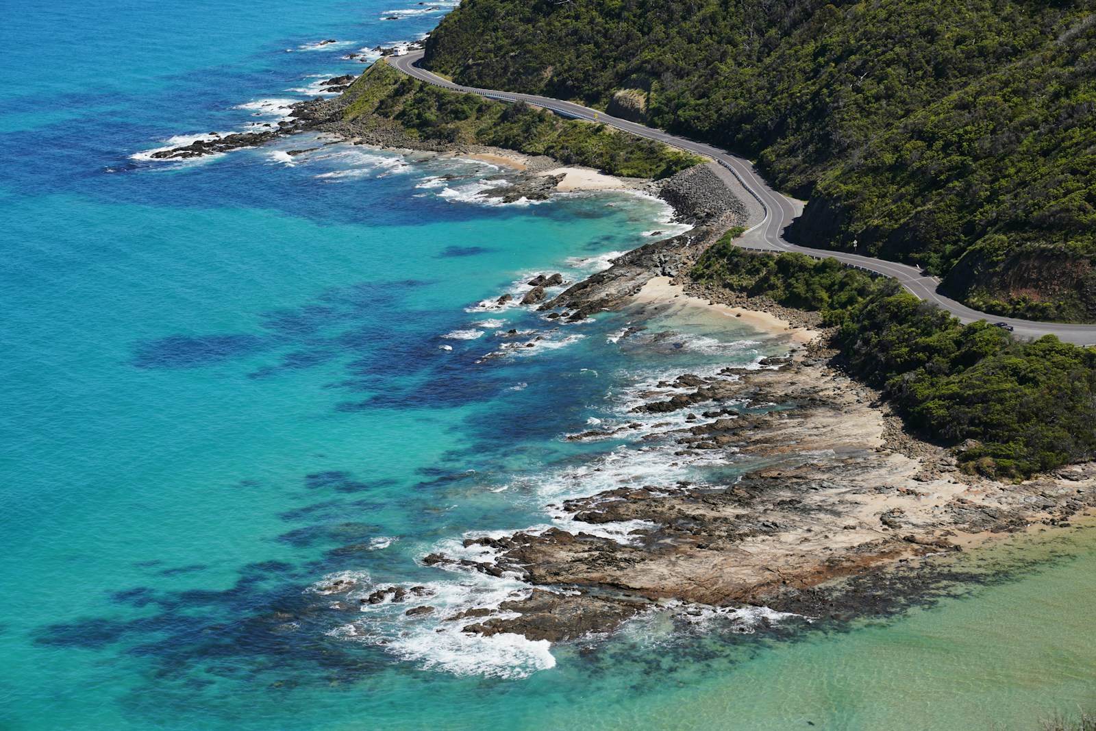 Aerial view of the Great Ocean Road winding along the Surf Coast near Lorne — teal water, rock shelves and coastal scrub.