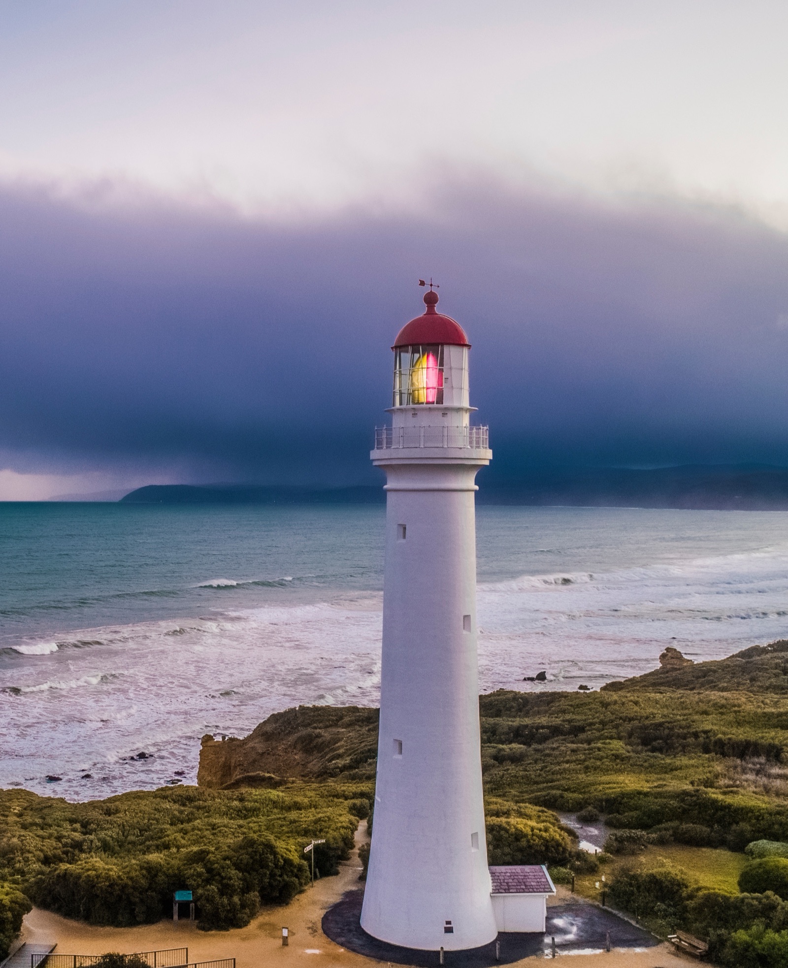 Split Point Lighthouse at dusk, Aireys Inlet — white tower, red cap, light on, as a southerly front rolls in over the Southern Ocean and surf breaks on the beach below.