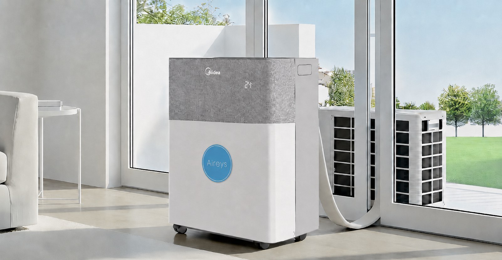 A Midea PortaSplit air conditioner wearing our roundel, standing on its castors in a bright living room beside patio doors. Its hose runs out through the open door to a compact outdoor unit sitting on the terrace — no bracket, nothing fixed to the building.